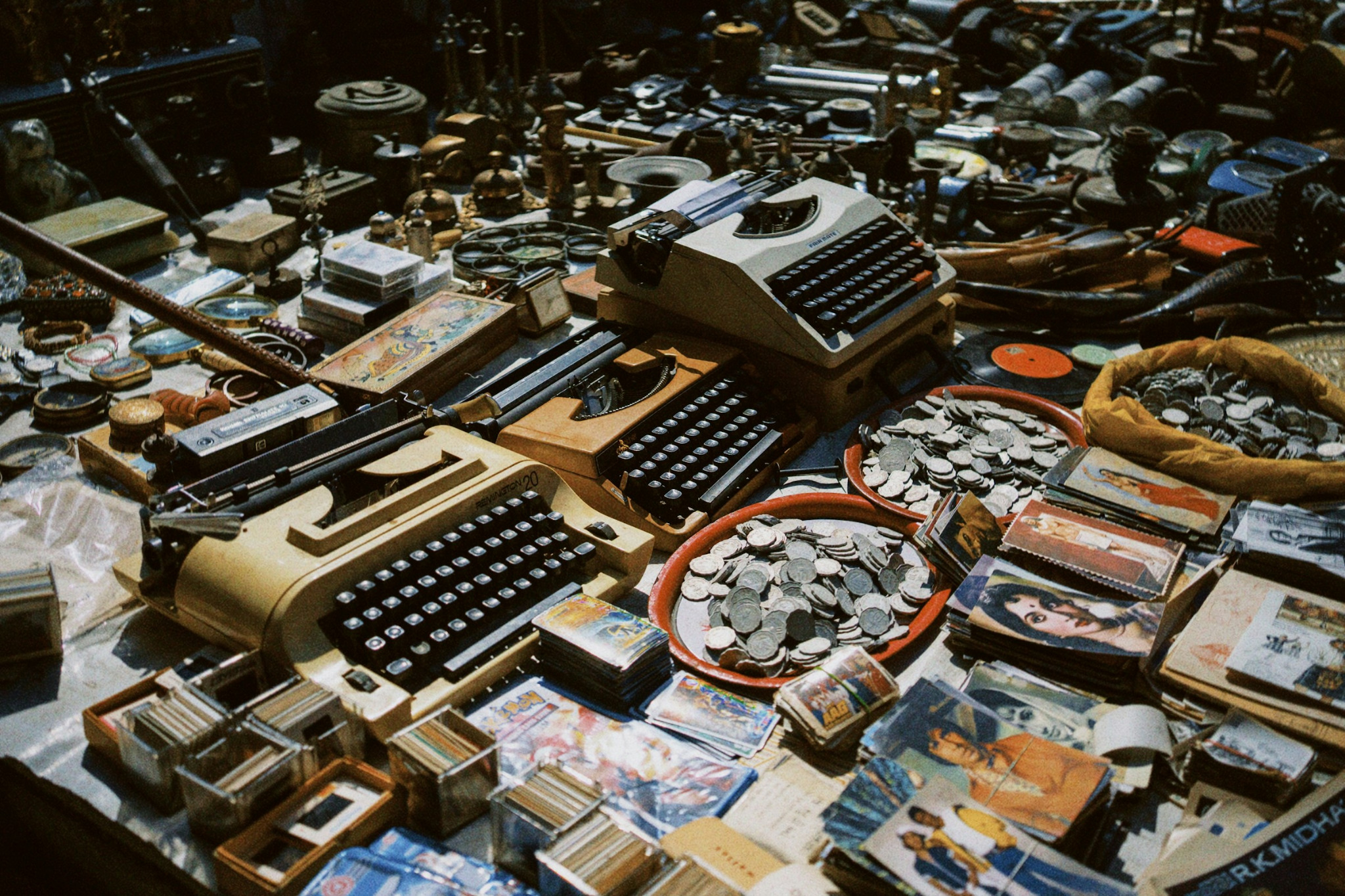 A crowded tabletop filled with vintage typewriters, records, photographs, cards, books, and collected objects