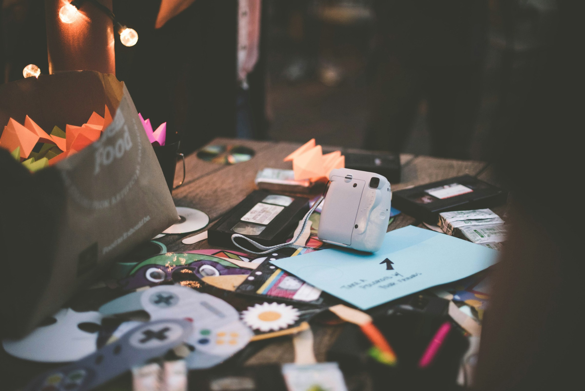A dimly lit table scattered with instant cameras, paper shapes, photographs, and art materials beneath warm string lights