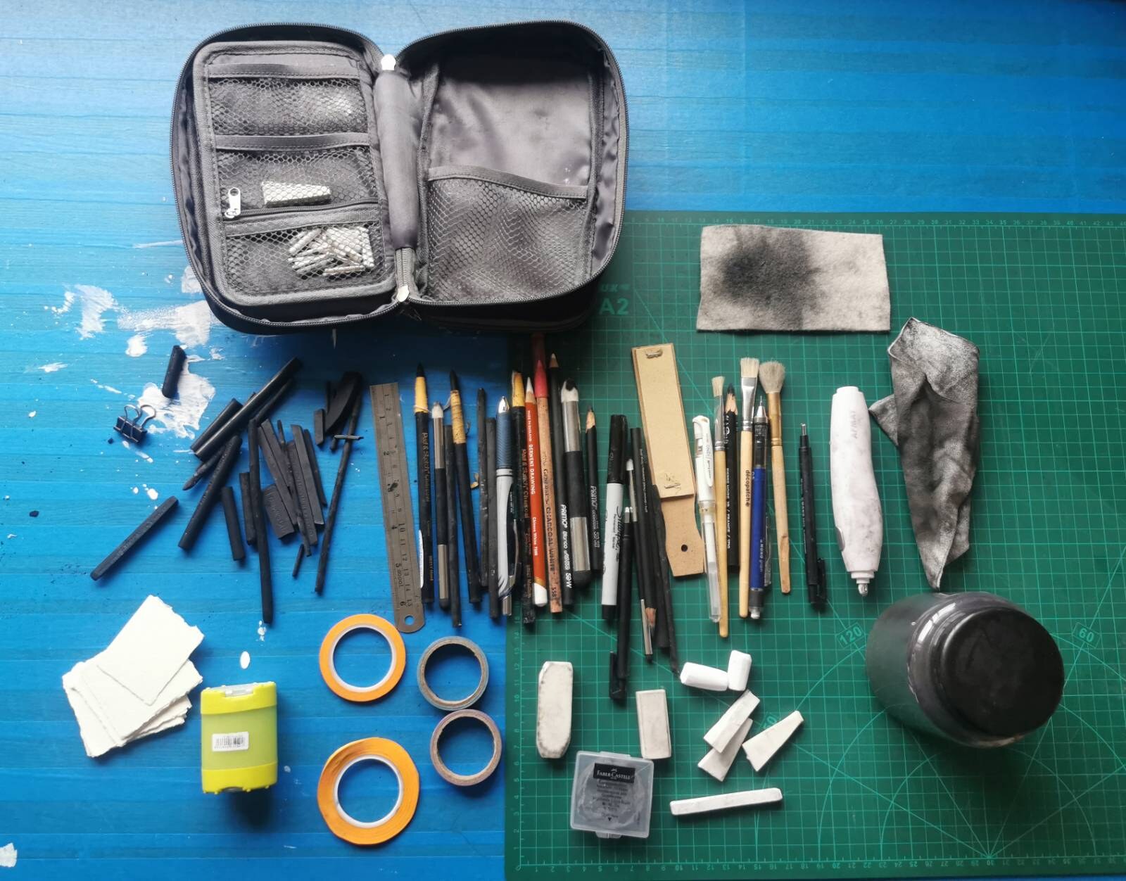 Charcoal sticks, pencils, brushes, erasers, tape, cloth, and scraps laid out on a blue table
