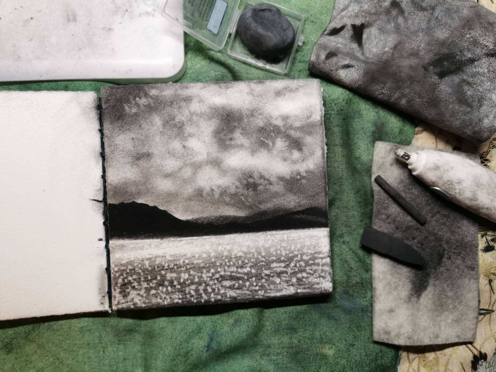 A small charcoal landscape study with sparkling light moving across water, surrounded by working materials