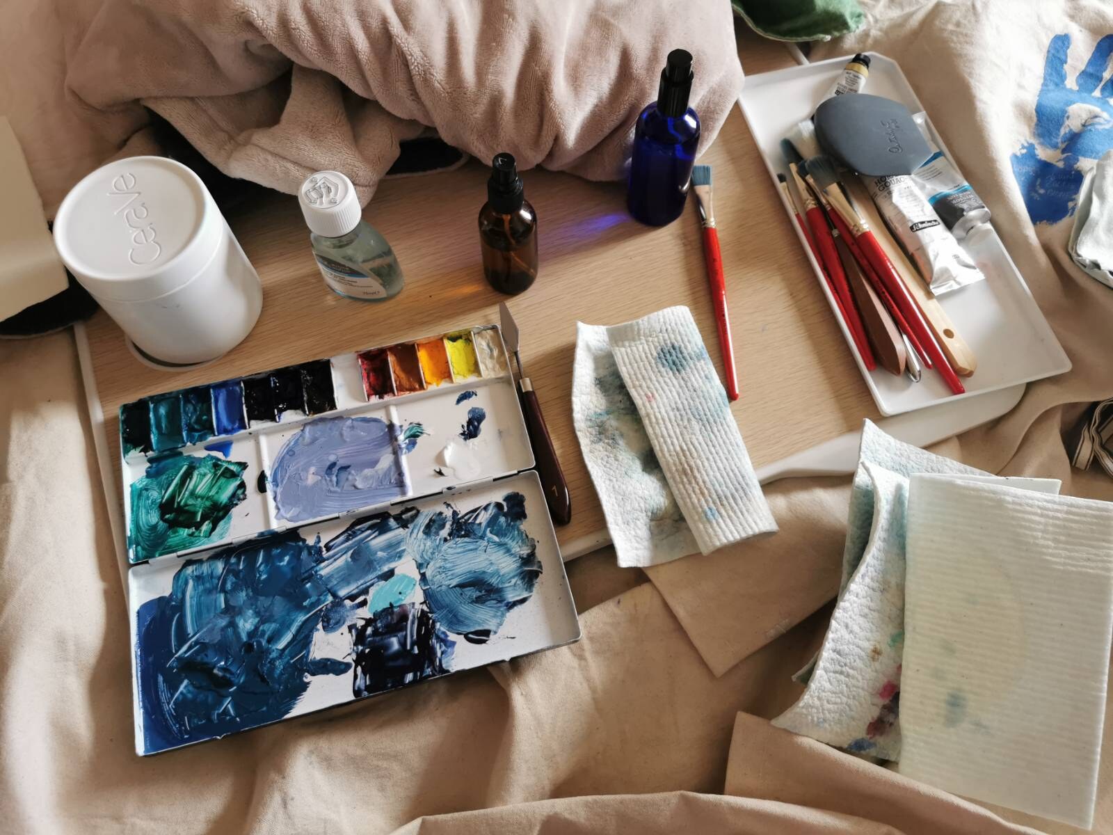 Water-media palette, brushes, bottles, cloths, and tools arranged on a tray and blankets in bed